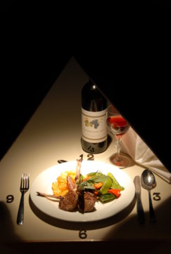 Ein Teller mit Lammkoteletts, Gemüse und Kartoffeln steht auf einem Tisch in Form einer Uhr, umgeben von Besteck, mit einer Flasche Wein und einem Glas Rotwein.