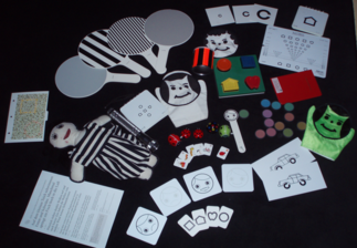 Eine Sammlung von visuellen und taktilen Hilfsmitteln zur Seh- und Wahrnehmungsförderung, darunter kontrastreiche Karten, bunte Formen und Puppen auf schwarzem Hintergrund.