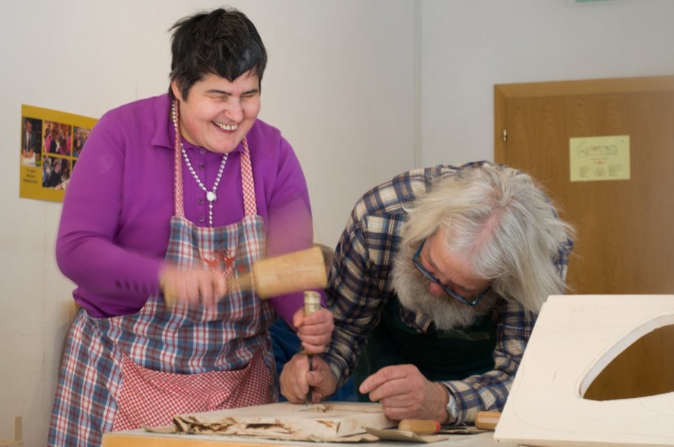 Eine lächelnde Frau und ein älterer Mann arbeiten zusammen an einem Holzprojekt, beide halten Werkzeuge in der Hand.