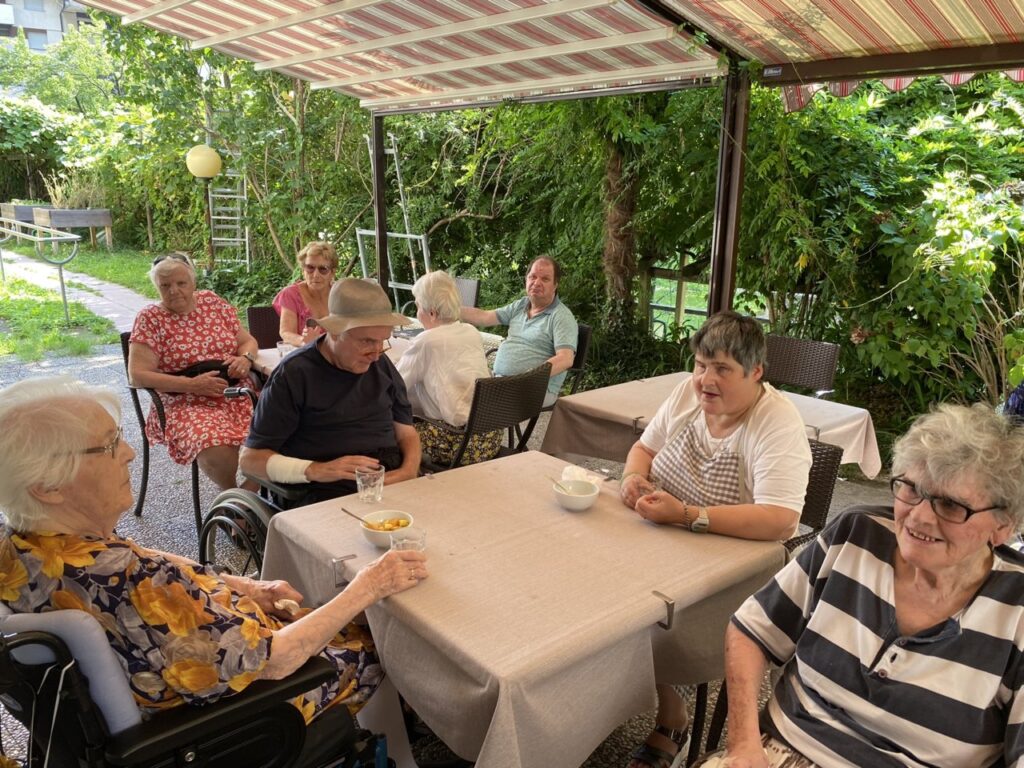 Un gruppo di anziani è seduto ai tavoli all'aperto, all'ombra di una tettoia, e chiacchiera in un giardino verdeggiante.