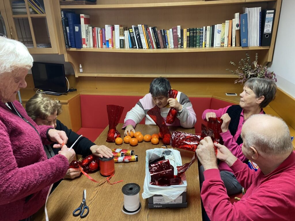 Un gruppo di anziani è seduto attorno a un tavolo e sta incartando regali e mandarini con carta rossa in una stanza con una libreria.