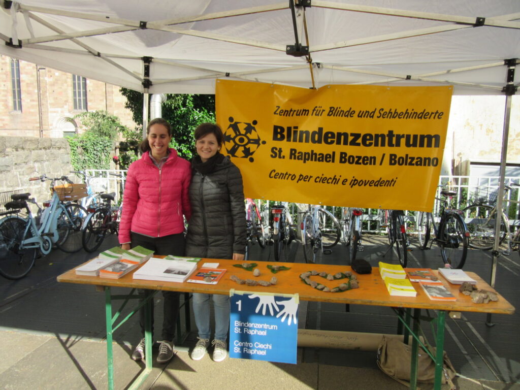 Due persone sono in piedi presso uno stand informativo del Centro Ciechi San Raphael di Bolzano, sotto un gazebo bianco circondato da biciclette.