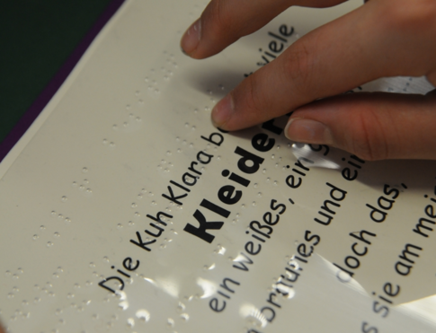 Eine Hand liest unter einem Text in Schwarzschrift einen Text in Brailleschrift.