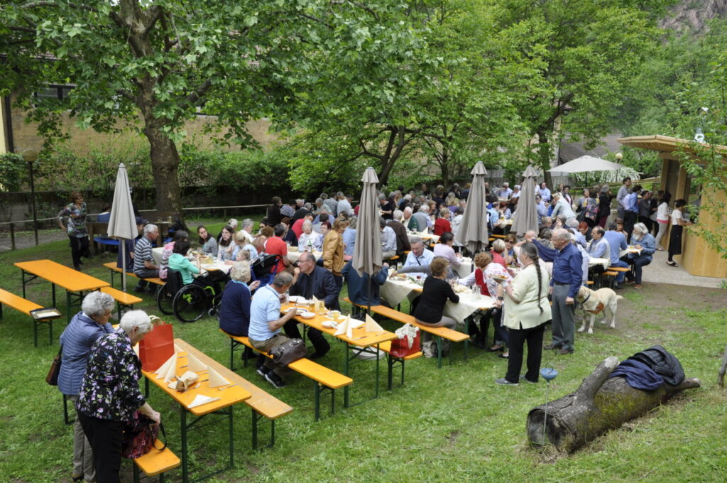 Eine große Gruppe von Menschen sitzt an langen Tischen in einem grünen Park, darunter Menschen im Rollstuhl und ein Blindenhund, während sie gemeinsam ein Fest im Freien genießen.
