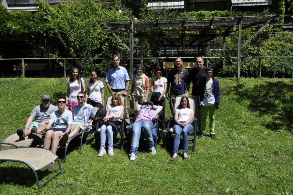 Un gruppo di persone è seduto e in piedi su un prato verde davanti a uno scenario naturale.
