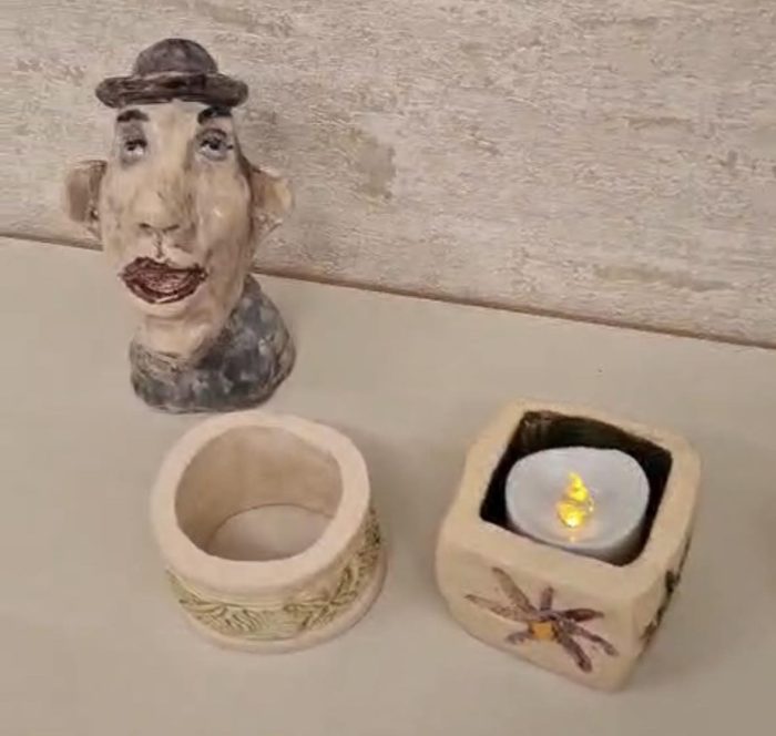 Drei Deko-Objekte auf einem Tisch: ein Keramikkopf, ein runder Behälter und ein Kerzenhalter mit brennender Kerze.