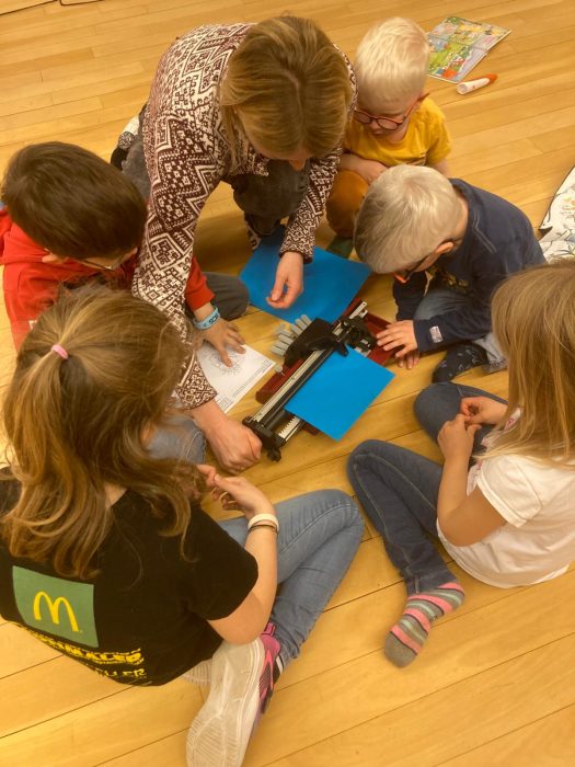 Eine Expertin sitzt mit mehreren Kindern auf dem Boden und zeigt ihnen eine Braille-Schreibmaschine. Die Kinder tasten und beobachten neugierig das Gerät und die Materialien.