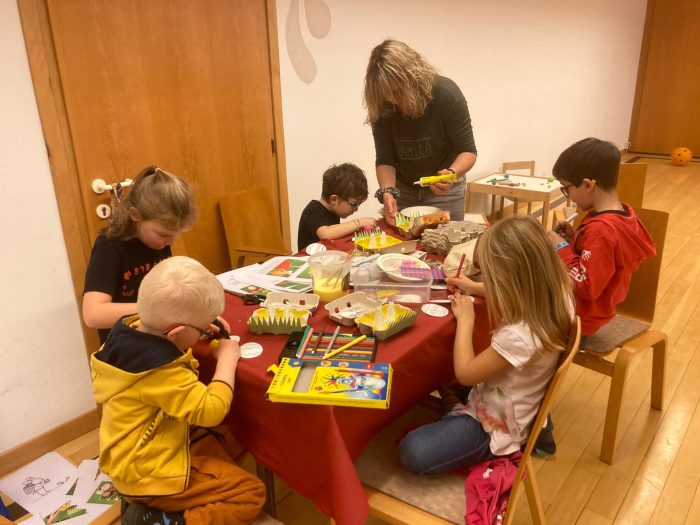 Mehrere Kinder sitzen an einem Tisch und basteln gemeinsam mit einer Betreuerin. Auf dem Tisch liegen Farben, Papier und verschiedene Bastelmaterialien.
