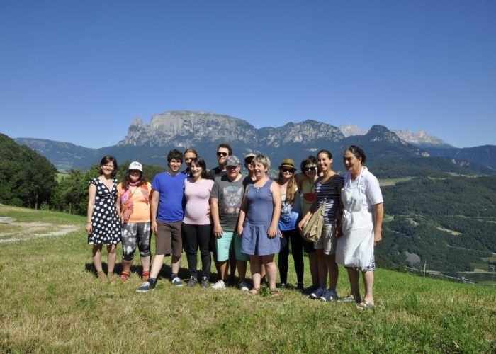 Eine Gruppe von Menschen steht auf einer Wiese vor einer Berglandschaft bei sonnigem Wetter.